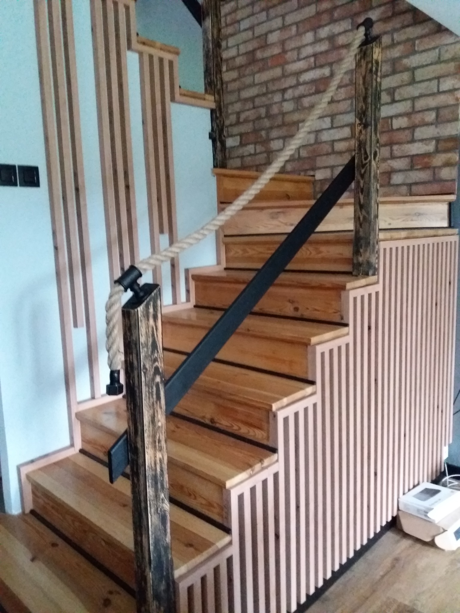 Drewniane schody z linową balustradą i pionowymi listwami na ścianie, wykończone cegłą w tle.