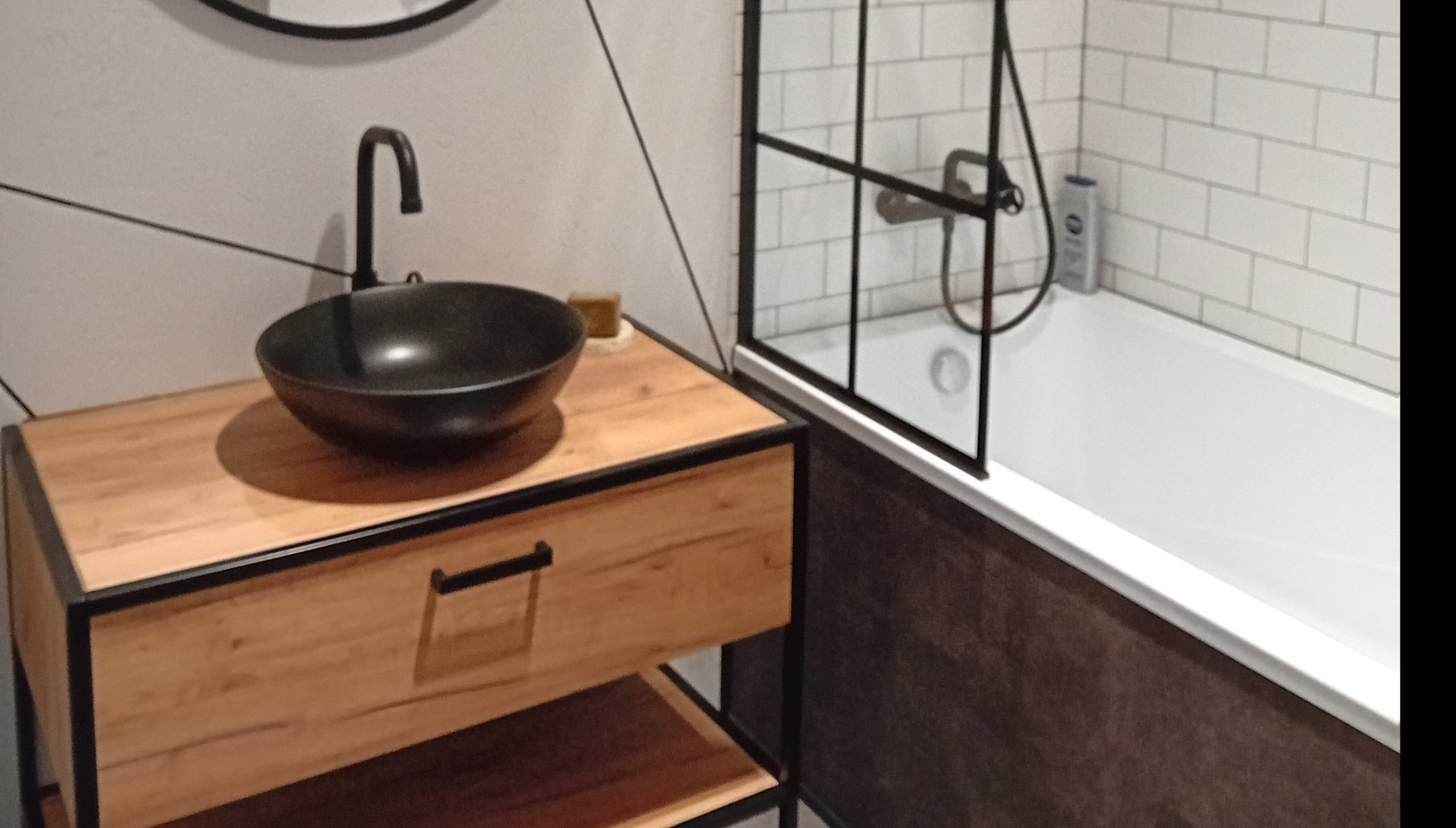 Nowoczesna łazienka z czarną umywalką nablatową na szafce z drewnianym frontem i czarnymi metalowymi elementami, obok kabina prysznicowa z białymi płytkami.