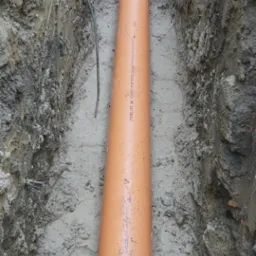 Pomarańczowa rura kanalizacyjna ułożona w wykopie w ziemi, Ostrów Wielkopolski. Widoczne ślady po wykopie i kabel leżący obok rury.