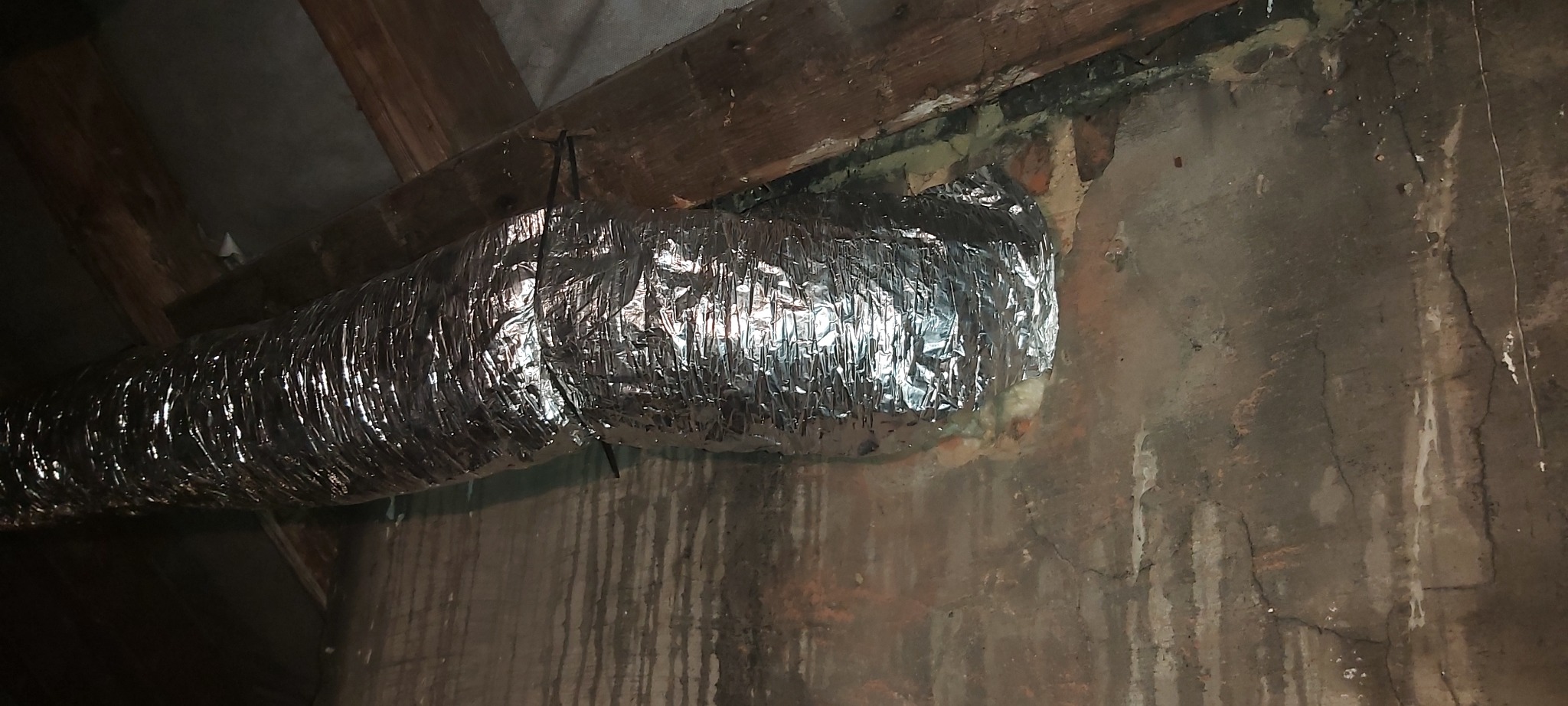 Elastyczny, aluminiowy przewód wentylacyjny przymocowany do drewnianej belki na tle szarej, wilgotnej ściany z widocznymi zaciekami.