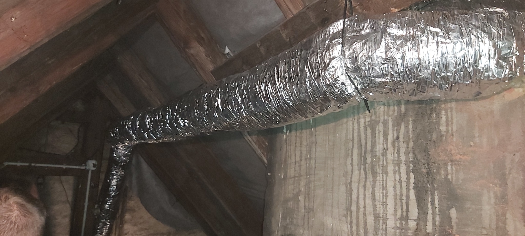 Elastyczny, izolowany przewód wentylacyjny aluminiowy zawieszony w przestrzeni poddasza z drewnianymi krokwiami i ścianą z zaciekami.