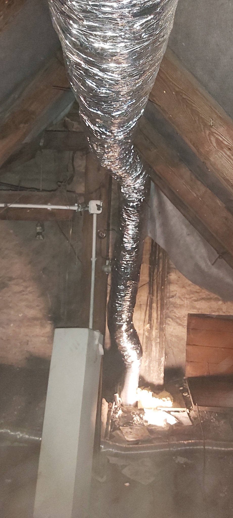 Elastyczny, srebrny przewód wentylacyjny zainstalowany w przestrzeni poddasza, widoczne drewniane elementy konstrukcyjne dachu oraz izolacja.