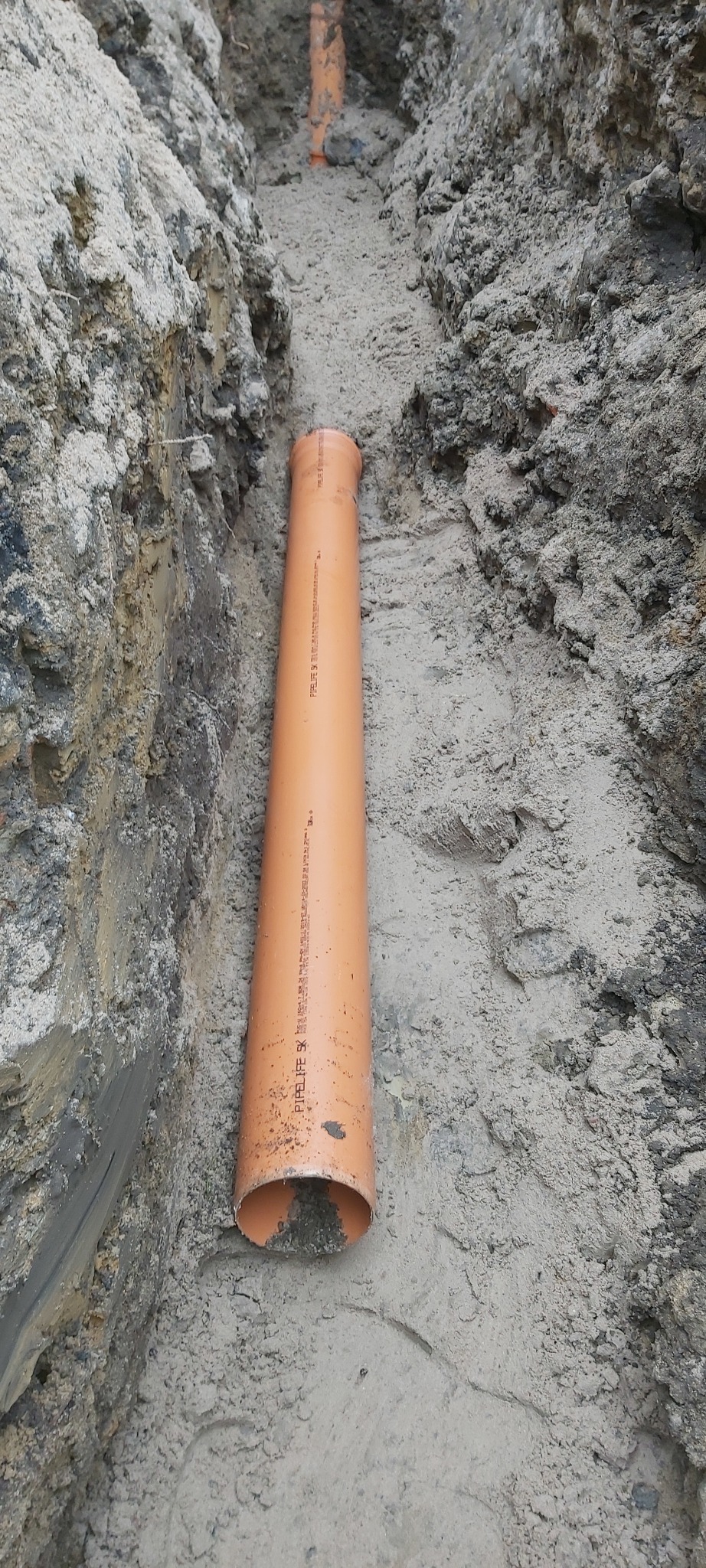 Pomarańczowa rura kanalizacyjna ułożona w wykopie ziemnym, widoczne ślady po pracy ziemnej.