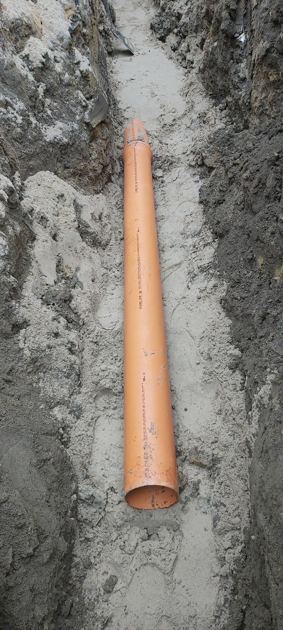 Pomarańczowa rura kanalizacyjna ułożona w wykopie ziemnym z piaskiem i gliną. Widoczne napisy na rurze.