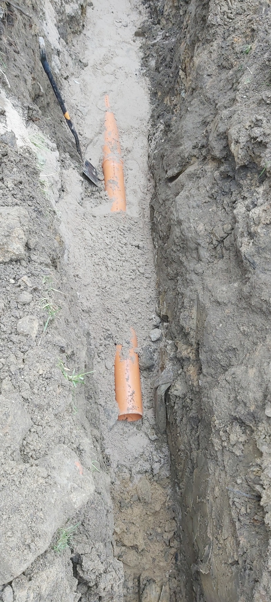 Wykop z ułożonymi pomarańczowymi rurami kanalizacyjnymi, widoczna łopata wbita w ziemię.