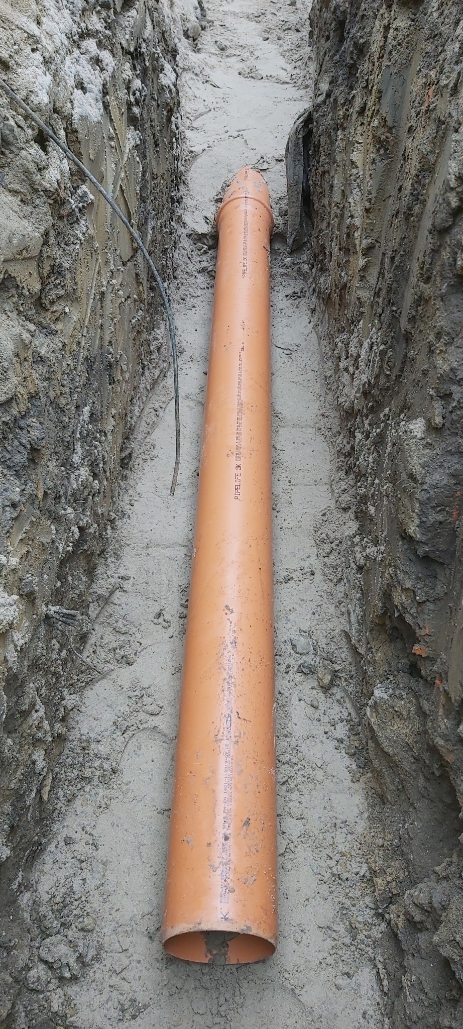 Pomarańczowa rura kanalizacyjna ułożona w wykopie w ziemi, Ostrów Wielkopolski. Widoczne ślady po wykopie i kabel leżący obok rury.