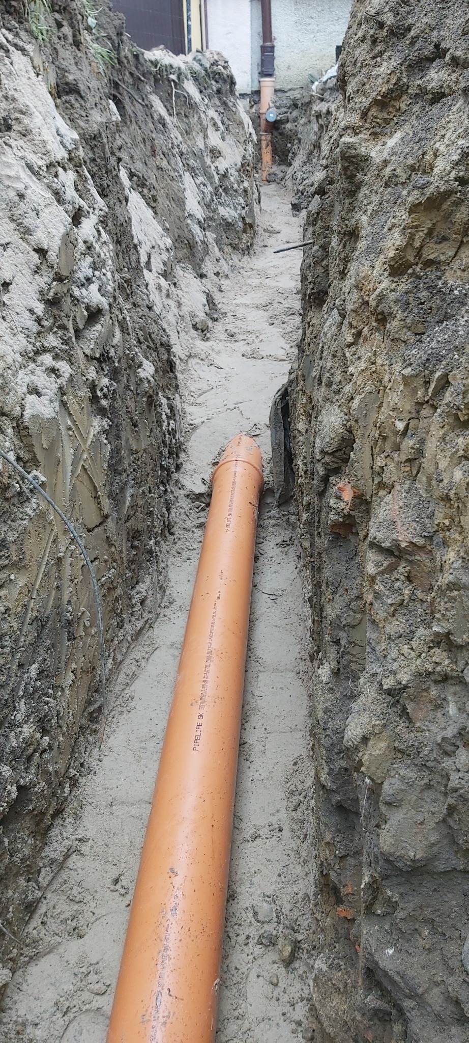 Pomarańczowa rura kanalizacyjna ułożona w wykopie ziemnym, widok z góry na dno wykopu.