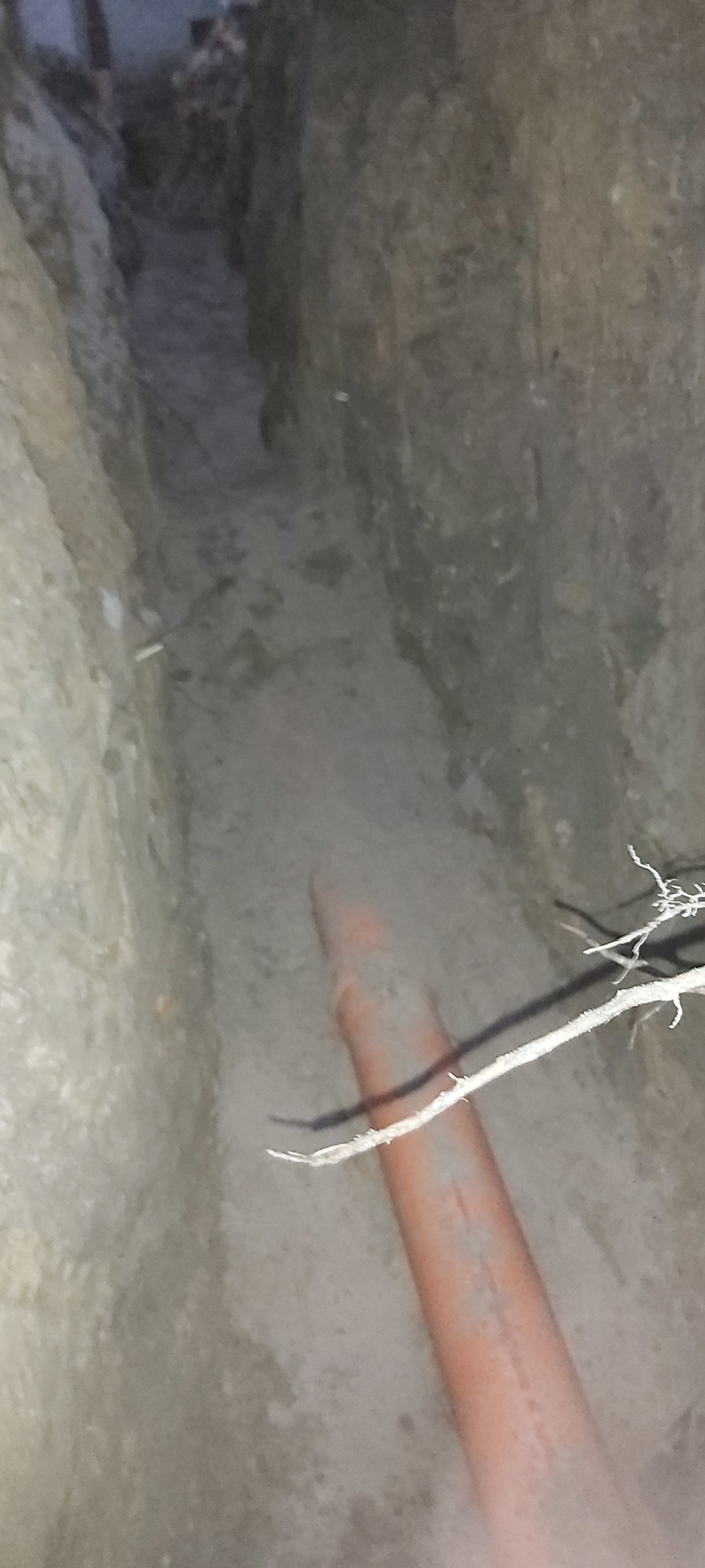 Pomarańczowa rura w wykopie ziemnym, widok z góry, oświetlenie słabe, fragment gałęzi w kadrze.