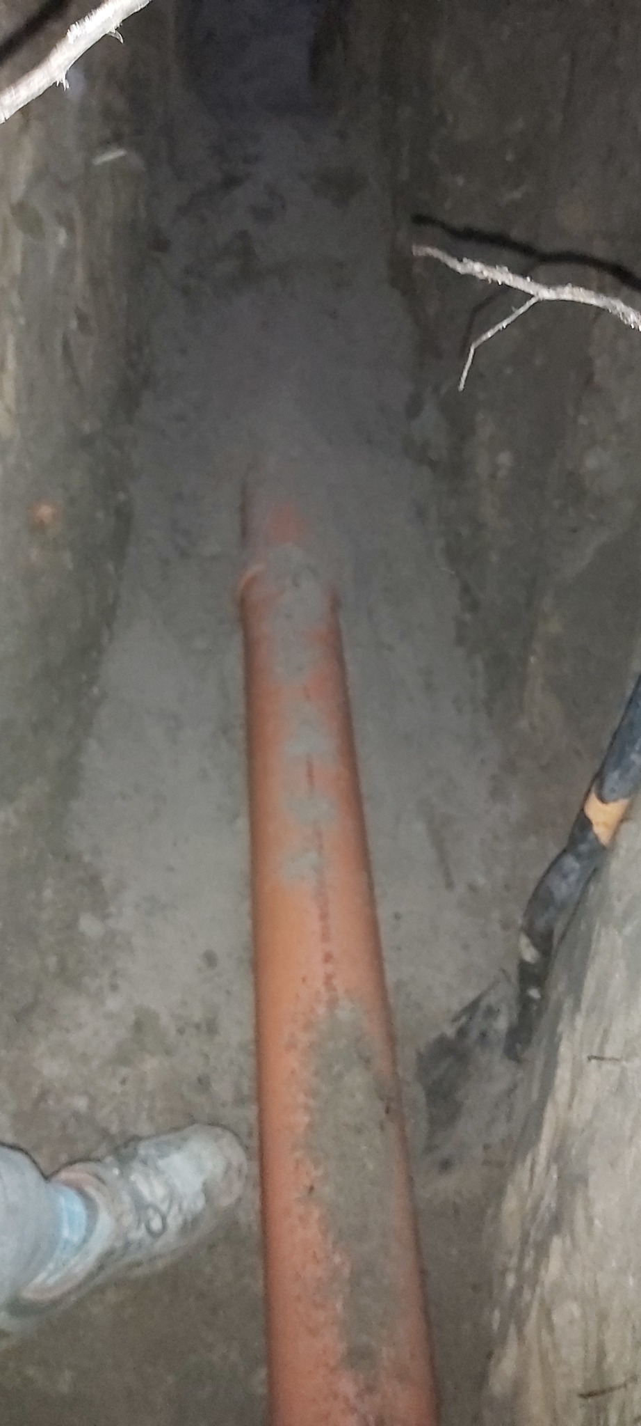 Pomarańczowa rura kanalizacyjna ułożona w wykopie ziemnym, widoczny fragment buta pracownika i kabel elektryczny.