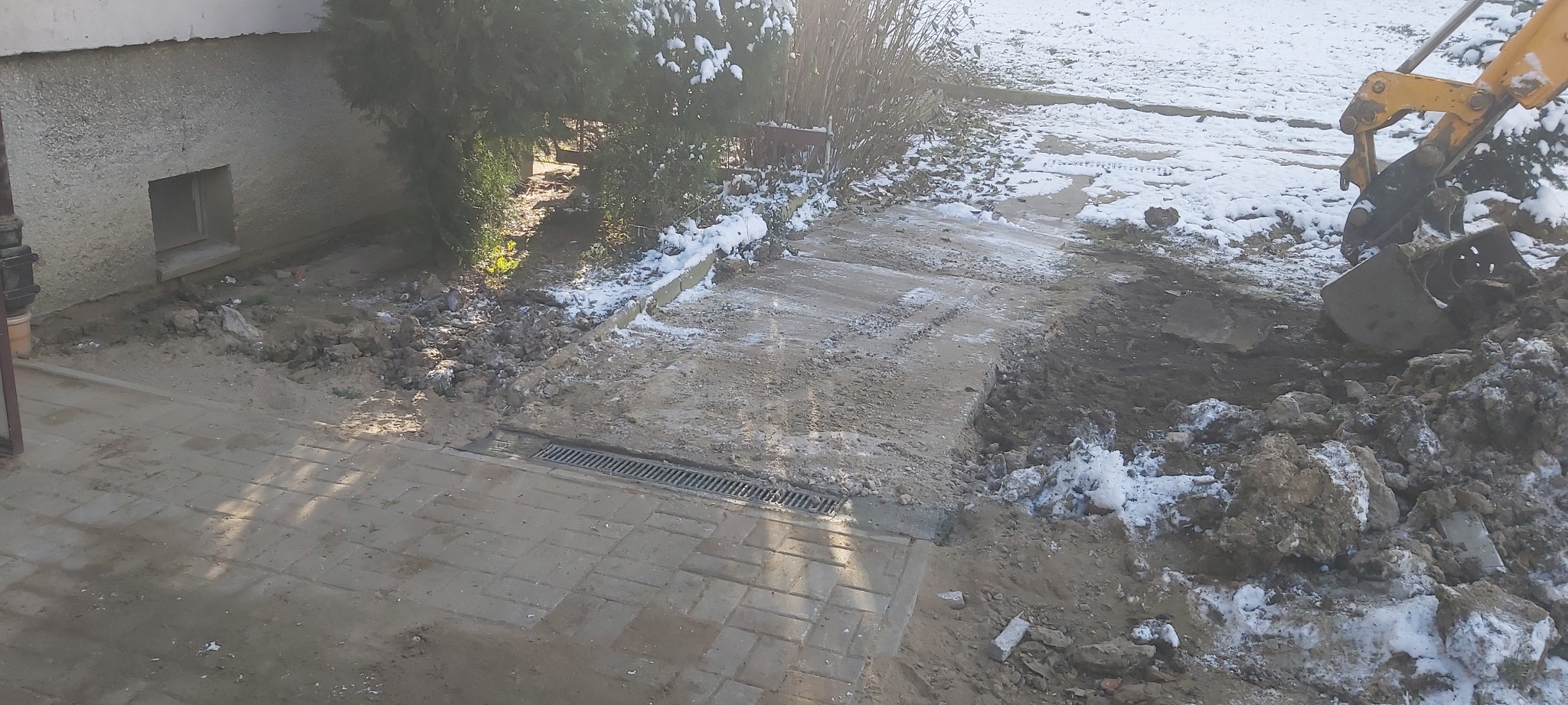 Żółta koparka podczas prac ziemnych przy budynku, widoczny fragment chodnika z kratką odpływową i resztki śniegu.