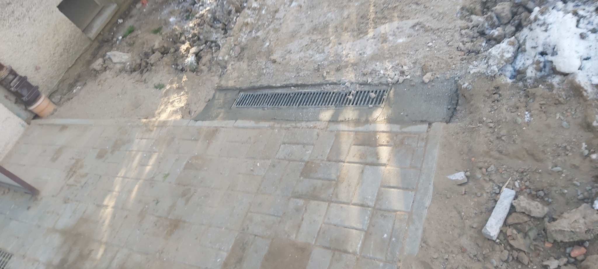 Zamontowana kratka odpływowa liniowa na betonowej podsypce, przylegająca do chodnika z kostki brukowej, teren wokół częściowo zasypany piaskiem i gruzem.