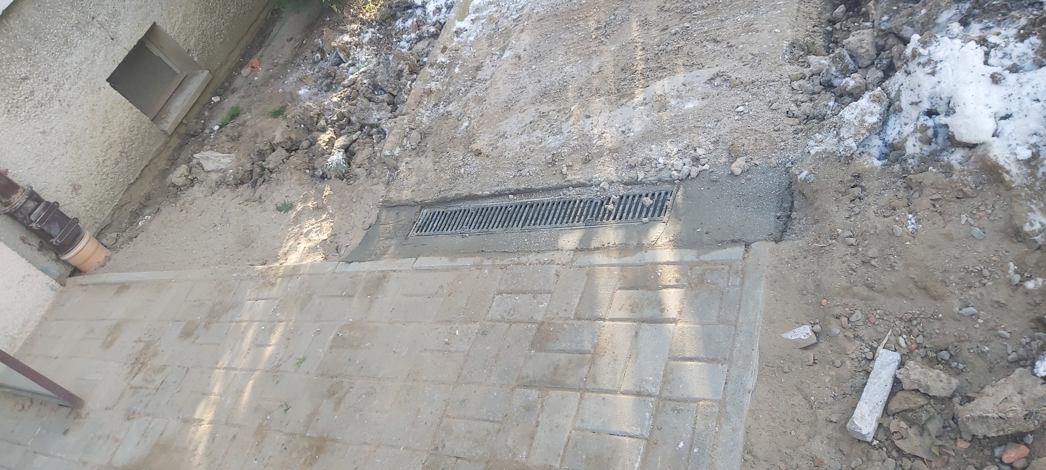 Ułożona kostka brukowa z zamontowaną kratką odpływową, widoczne ślady prac ziemnych i resztki śniegu.