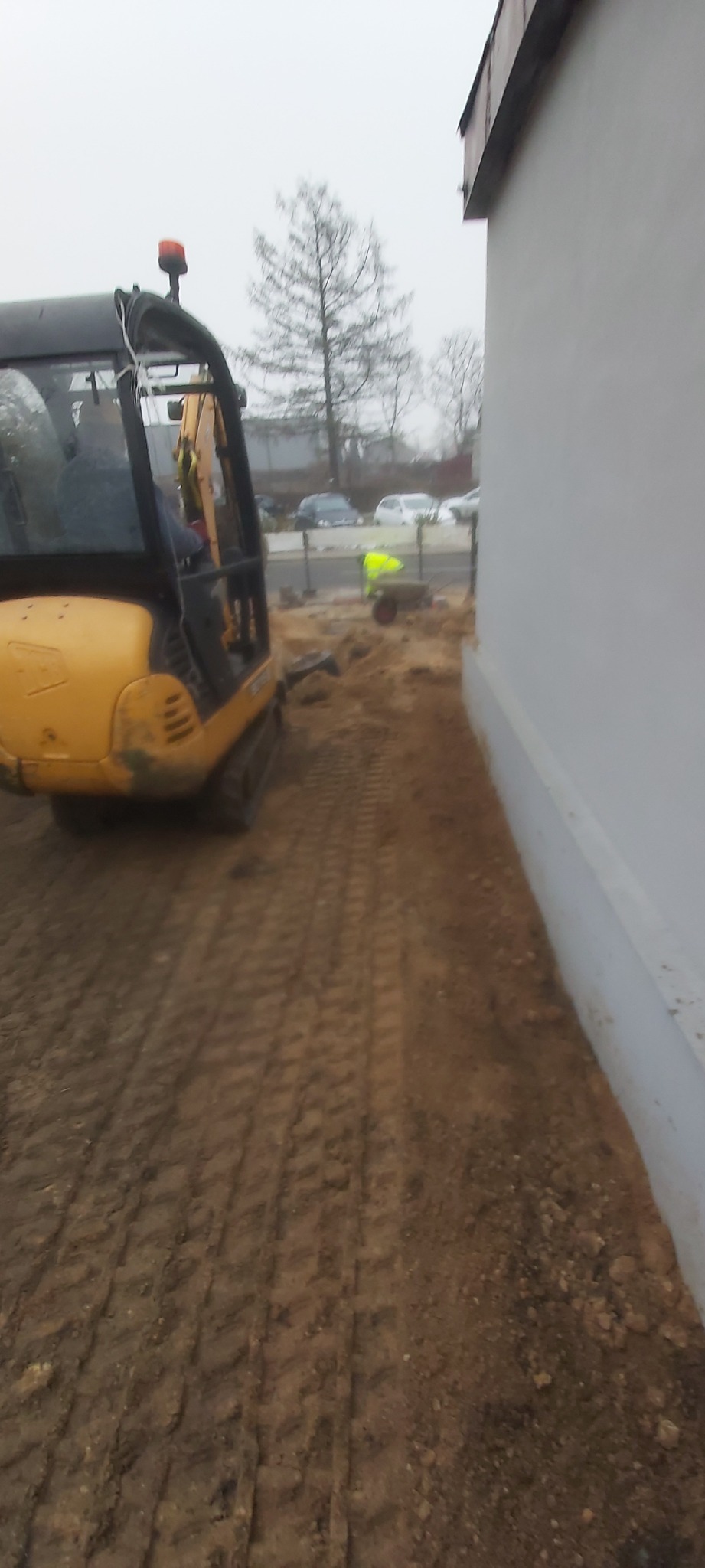Żółta mini koparka obok białego budynku, ślady opon na ziemi, w tle pracownik w żółtej kamizelce.