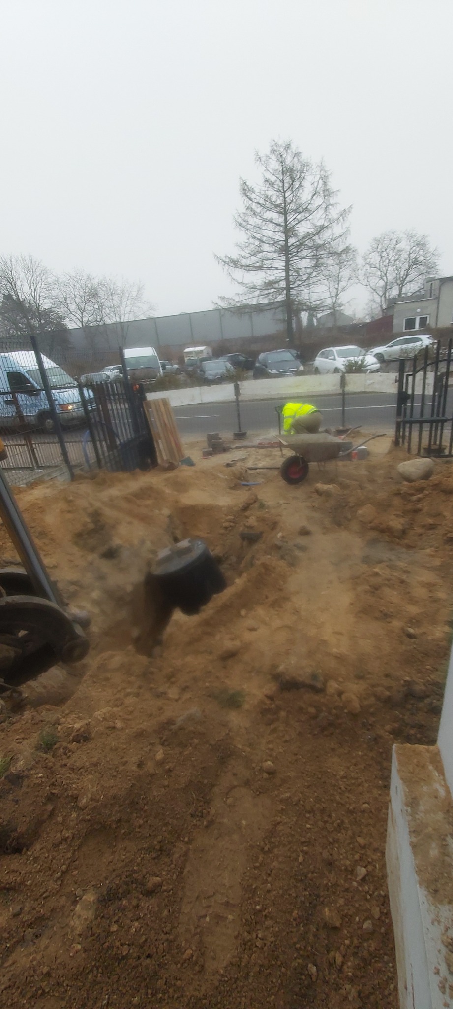 Koparka częściowo zagrzebana w ziemi, obok pracownik w kamizelce odblaskowej i taczka na tle ulicy i drzew