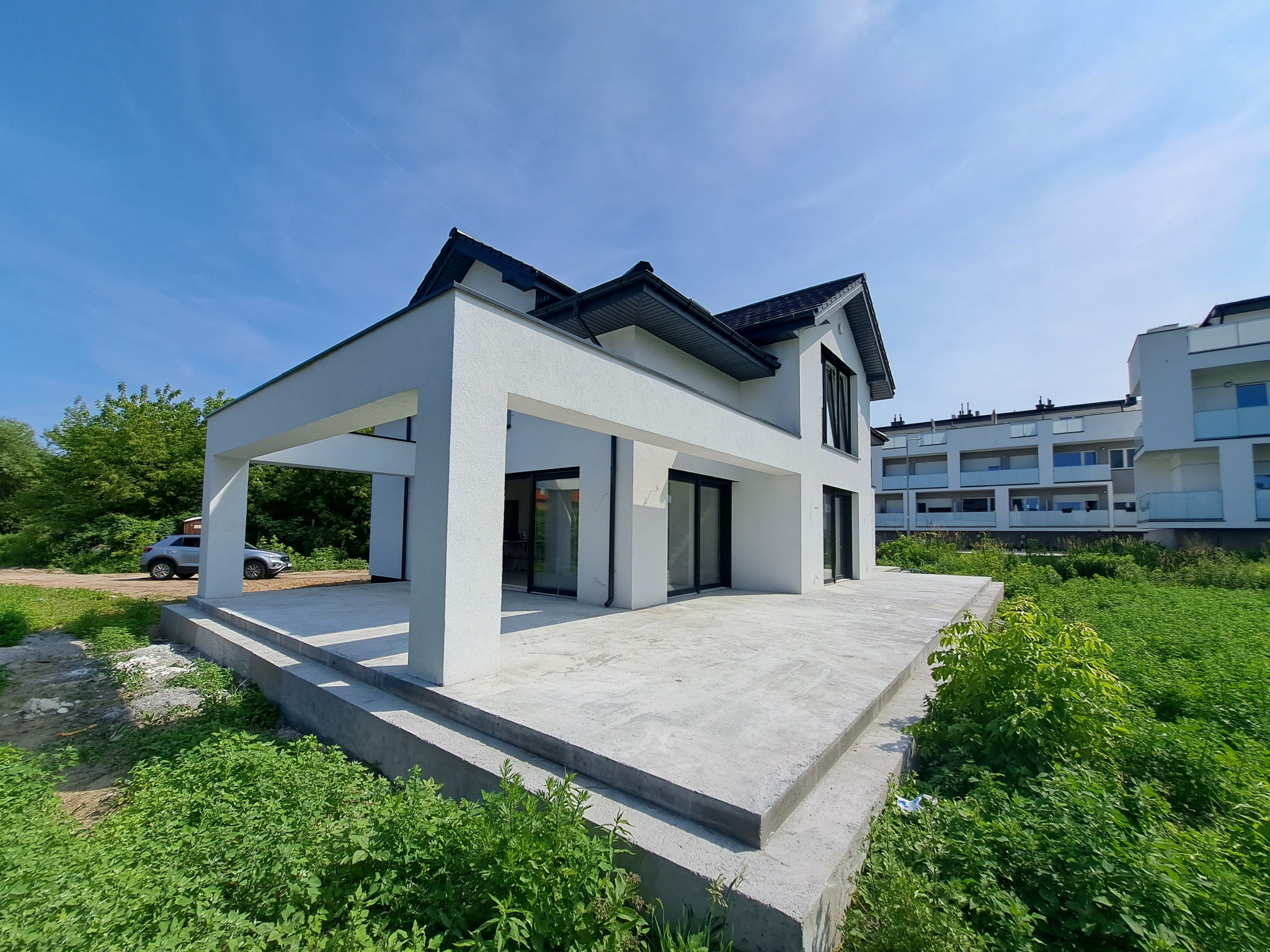 Nowoczesny, biały dom jednorodzinny z płaskim dachem i dużymi oknami, widok z niskiej perspektywy na tle błękitnego nieba i zieleni, z widocznym samochodem osobowym w tle.