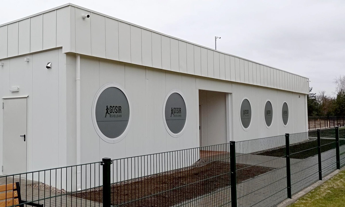 Biały budynek modułowy z okrągłymi oknami z logo, otoczony metalowym ogrodzeniem.