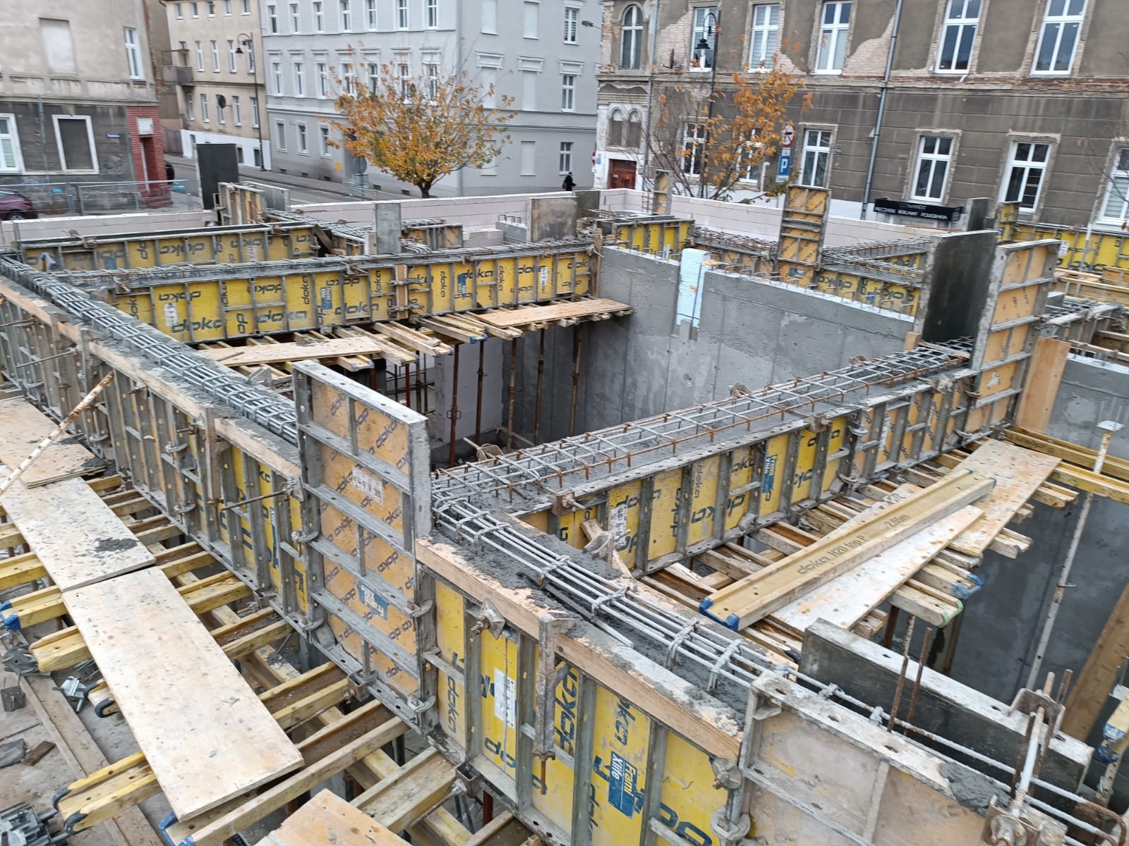 Fundamenty budynku w trakcie budowy z widocznymi szalunkami Doka i zbrojeniami, na tle miejskiej zabudowy.