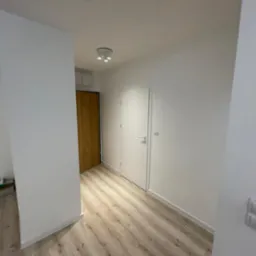 Wnętrze po remoncie: białe ściany, panele podłogowe w odcieniach drewna, widoczne dwie pary drzwi - drewniane i białe, oraz podw&oacute;jny włącznik światła na ścianie.