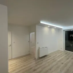 Jasne, wykończone mieszkanie z panelami podłogowymi, białymi ścianami, grzejnikiem i oknami balkonowymi, oświetlone listwą LED.