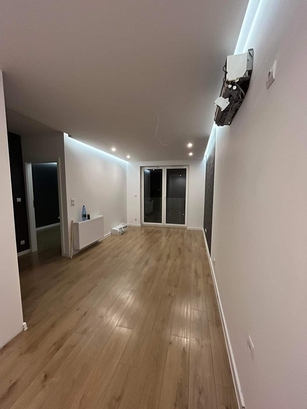 Nowoczesne wnętrze po remoncie: parkiet w odcieniach dębu, białe ściany z oświetleniem LED, widoczne drzwi balkonowe i niedokończony montaż klimatyzacji na ścianie.