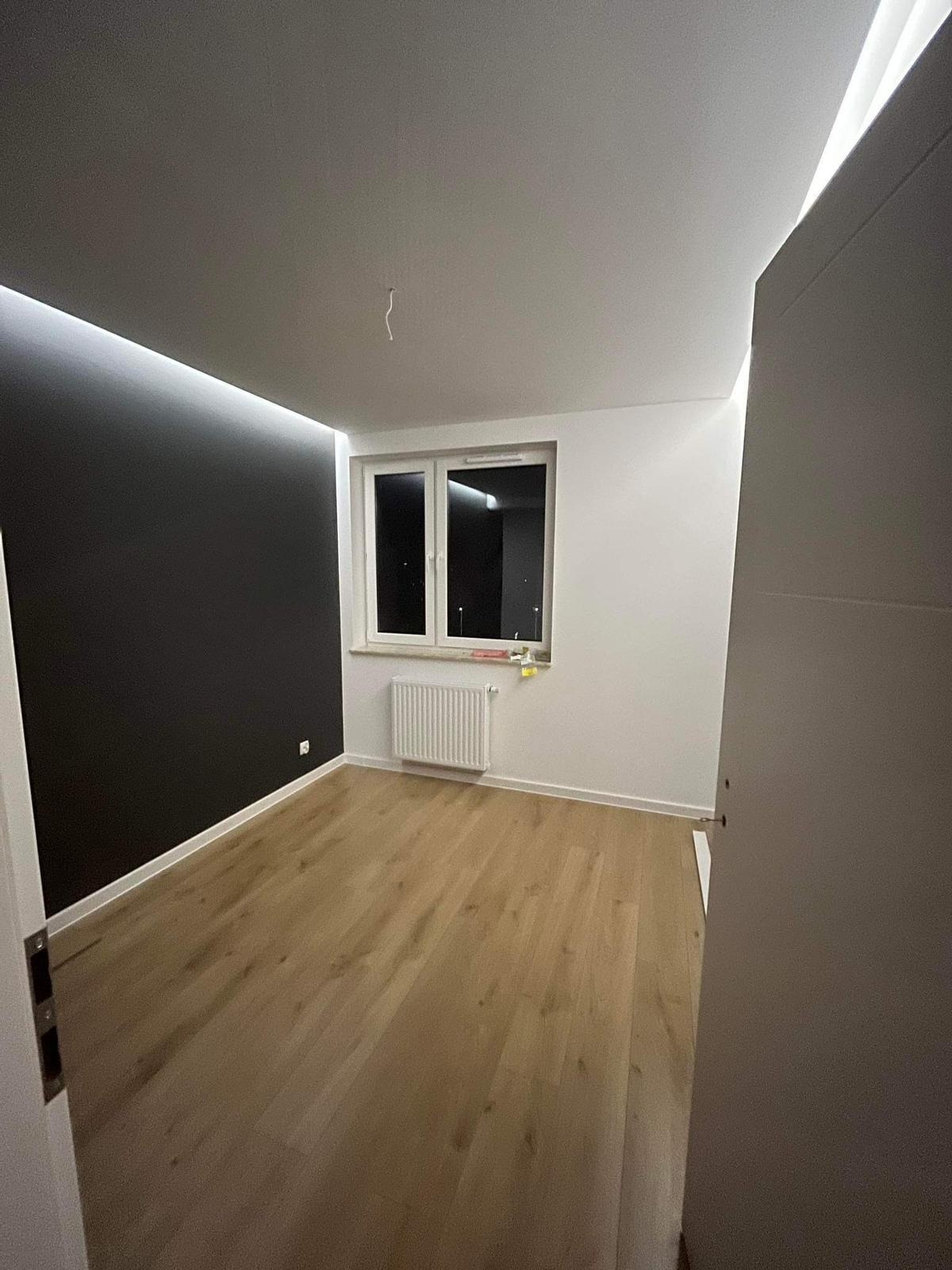 Wnętrze pokoju po remoncie: czarna ściana z oświetleniem LED na suficie, jasna podłoga, białe ściany z oknem i grzejnikiem.