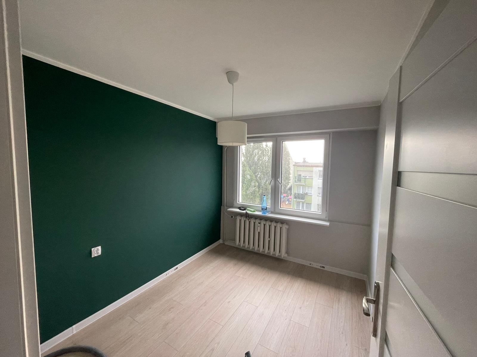 Pokój po remoncie: jedna ściana pomalowana na ciemny zielony kolor, druga na szaro, jasna podłoga imitująca drewno, białe listwy przypodłogowe, okno z białymi ramami, biały grzejnik, biała lampa...