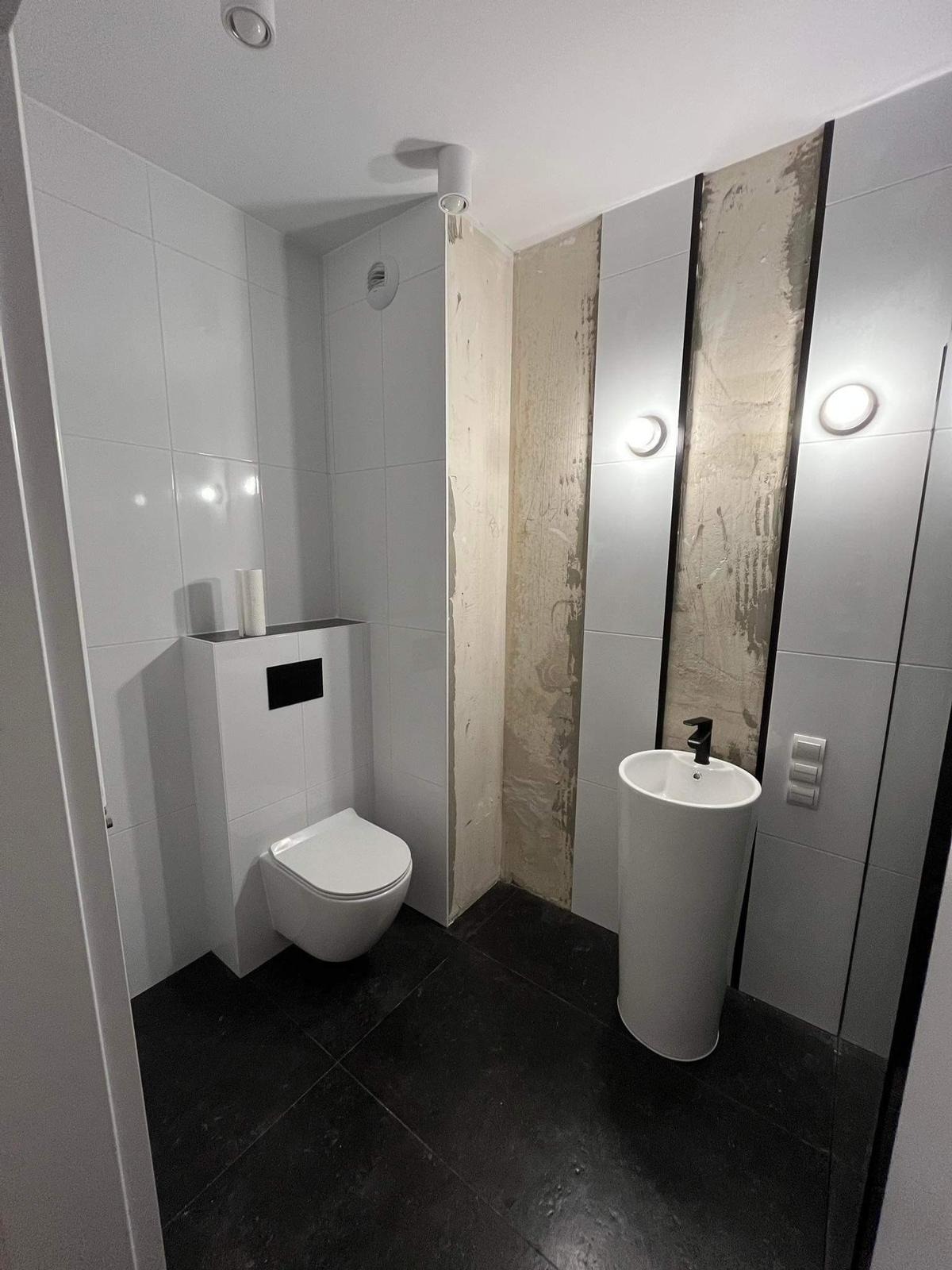Wnętrze łazienki w trakcie odnawiania, widoczna podwieszana toaleta, umywalka kolumnowa, czarne płytki podłogowe, białe płytki ścienne oraz fragment ściany w trakcie przygotowania pod wykończenie.