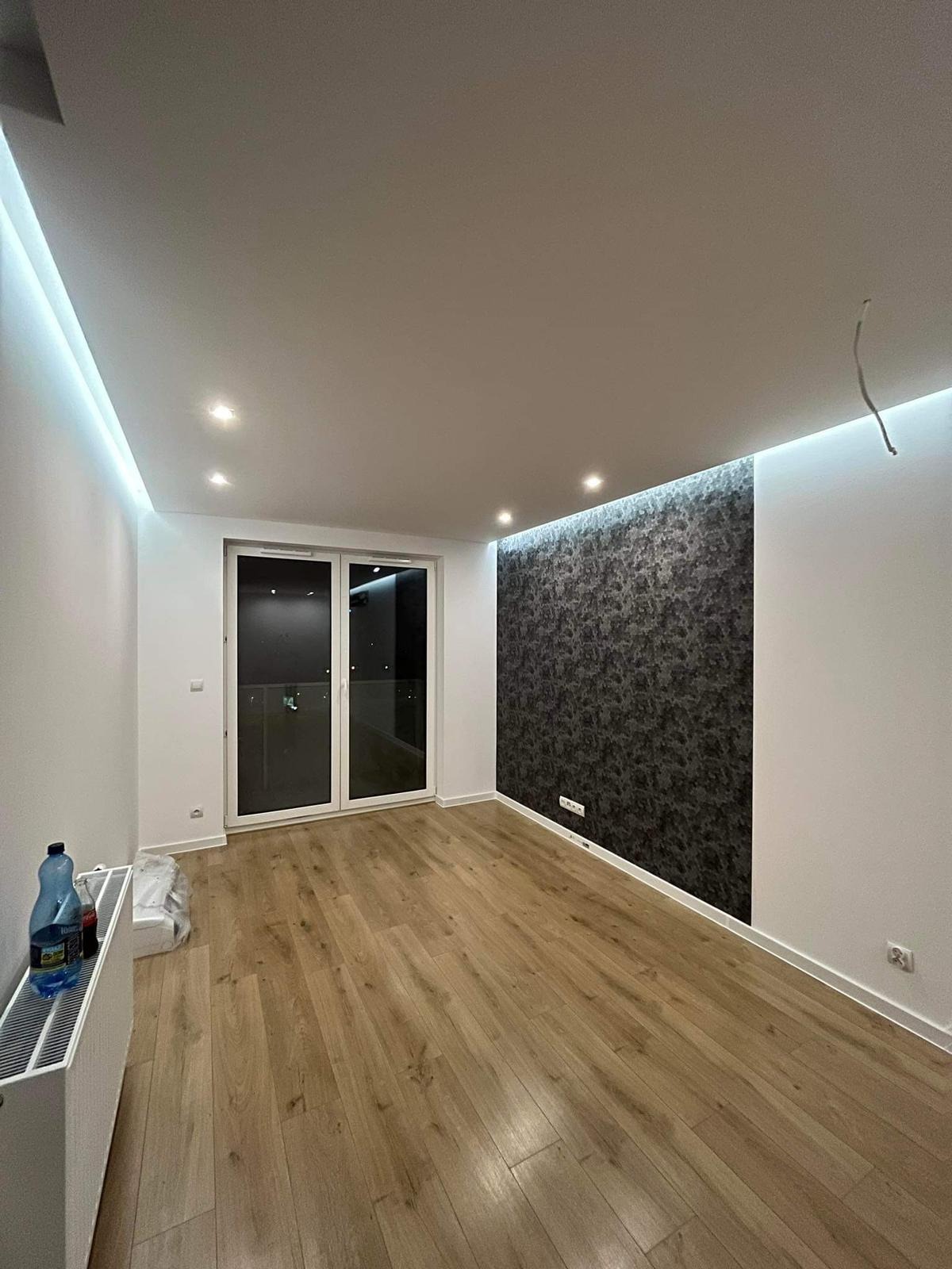 Nowoczesne wnętrze pokoju po remoncie: panele podłogowe w odcieniach drewna, ściany pomalowane na biało z fragmentem pokrytym tapetą o ciemnym, abstrakcyjnym wzorze, przesuwne drzwi balkonowe...