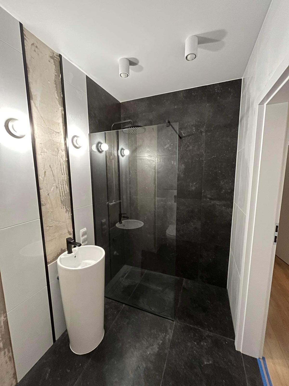Nowoczesna łazienka z kabiną prysznicową typu walk-in, czarną armaturą i umywalką wolnostojącą; widoczne fragmenty niedokończonych prac glazurniczych.