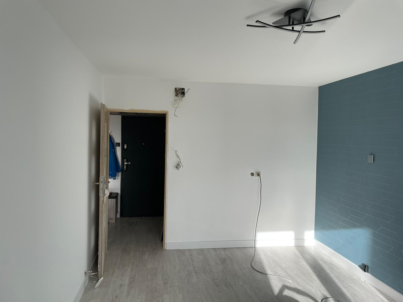 Pokój w trakcie remontu: otwarte drzwi wejściowe, niebieska kurtka wisząca w przedpokoju, ściana z niebieską tapetą w geometryczne wzory, gładkie białe ściany z wystającymi kablami, szara podłoga...