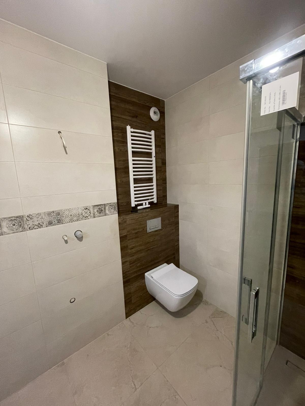 Nowoczesna łazienka po remoncie: białe i beżowe płytki, podwieszana toaleta, grzejnik drabinkowy na brązowej ścianie, kabina prysznicowa ze srebrnymi okuciami. Częściowo widoczne niedoróbki...