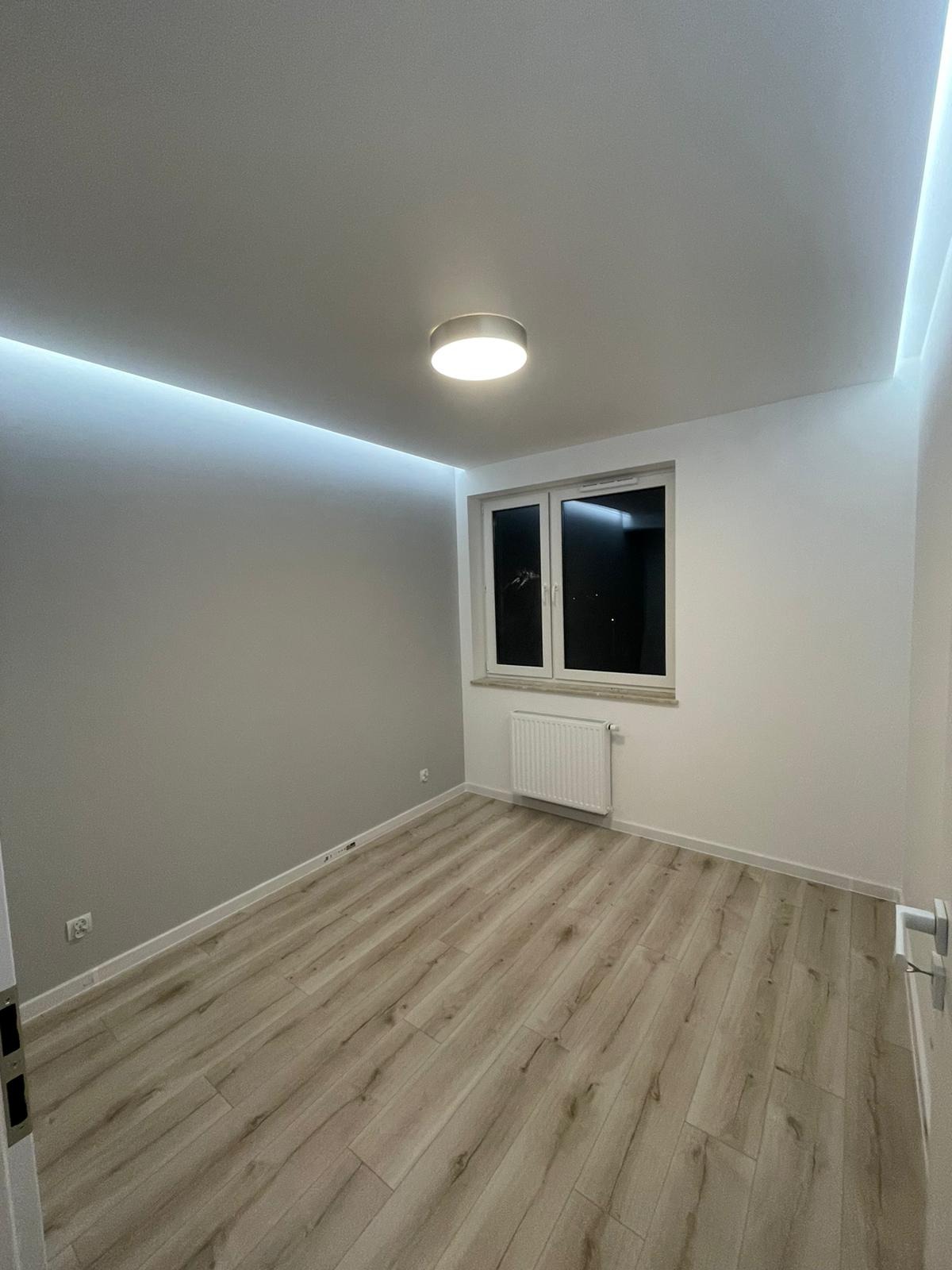 Wnętrze pokoju po remoncie: panele podłogowe w odcieniach drewna, szara ściana z listwą LED pod sufitem, biała ściana z oknem i grzejnikiem.