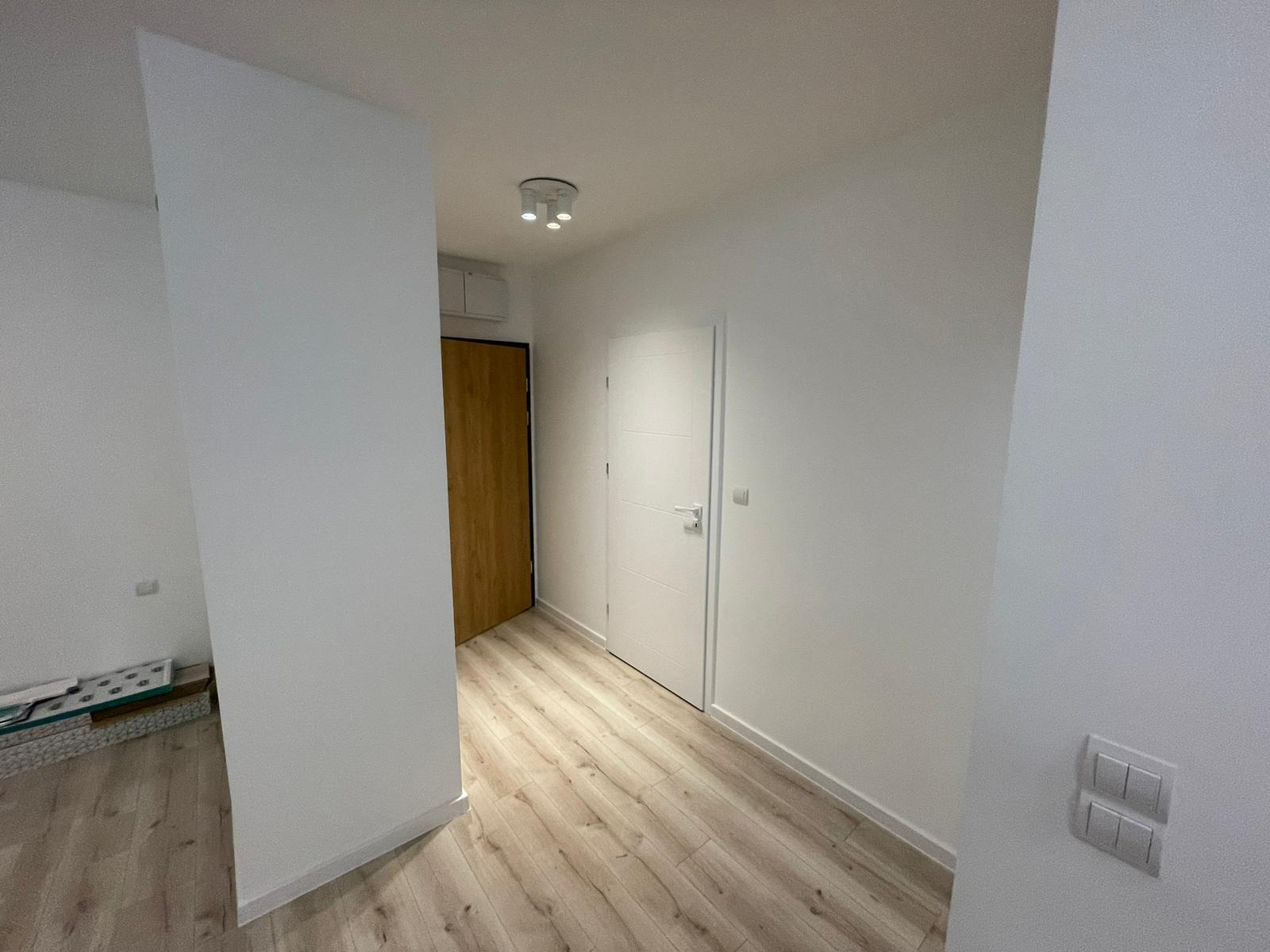 Wnętrze po remoncie: białe ściany, panele podłogowe w odcieniach drewna, widoczne dwie pary drzwi - drewniane i białe, oraz podwójny włącznik światła na ścianie.