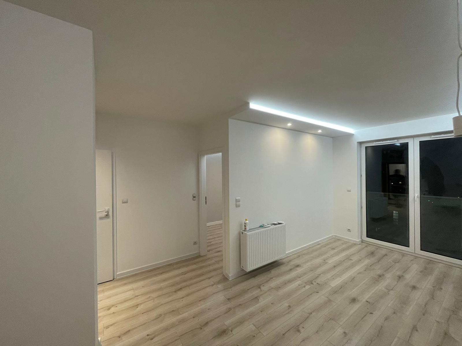 Jasne, wykończone mieszkanie z panelami podłogowymi, białymi ścianami, grzejnikiem i oknami balkonowymi, oświetlone listwą LED.