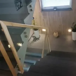 Nowoczesne schody z ciemnego kamienia z drewnianą poręczą i szklaną balustradą, oświetlone punktowymi lampami w ścianie, z roślinami doniczkowymi i dekoracyjnym koszykiem w tle.