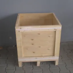 Pusta, jasna, drewniana skrzynia paletowa z czterema nogami, zabezpieczona taśmą, stojąca na szarej kostce brukowej przed szarą ścianą.