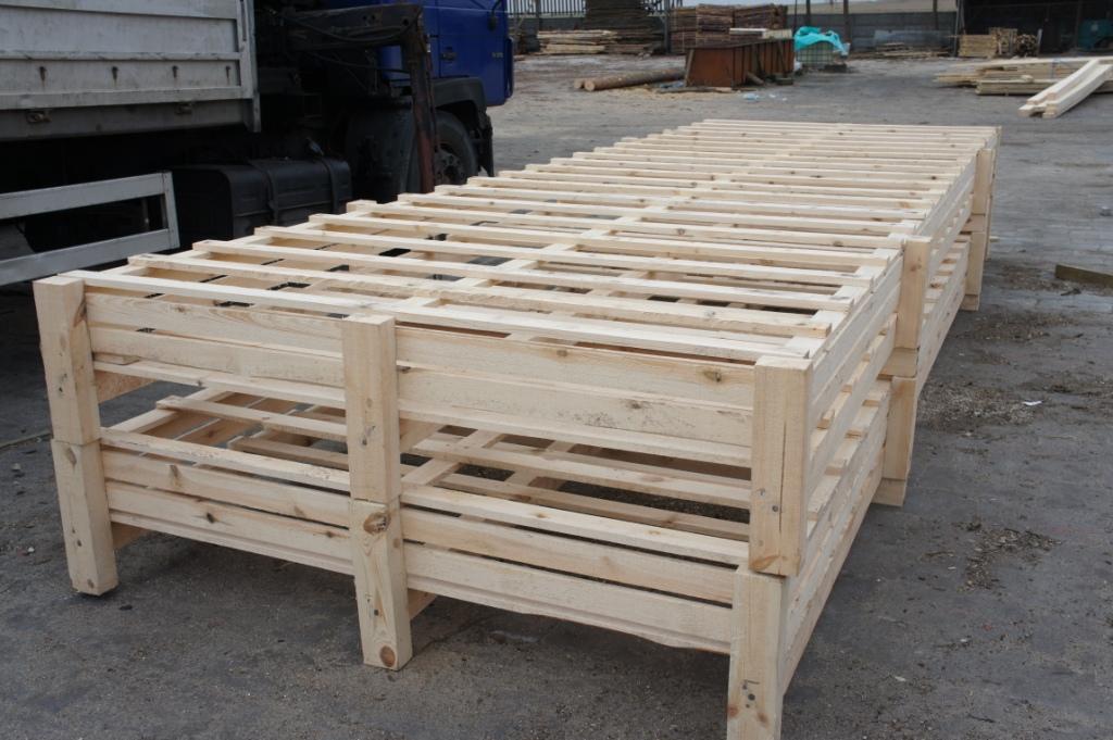 Stos jasnych, drewnianych skrzyń transportowych na paletach, ustawionych na zewnątrz, z fragmentem niebieskiej ciężarówki w tle.