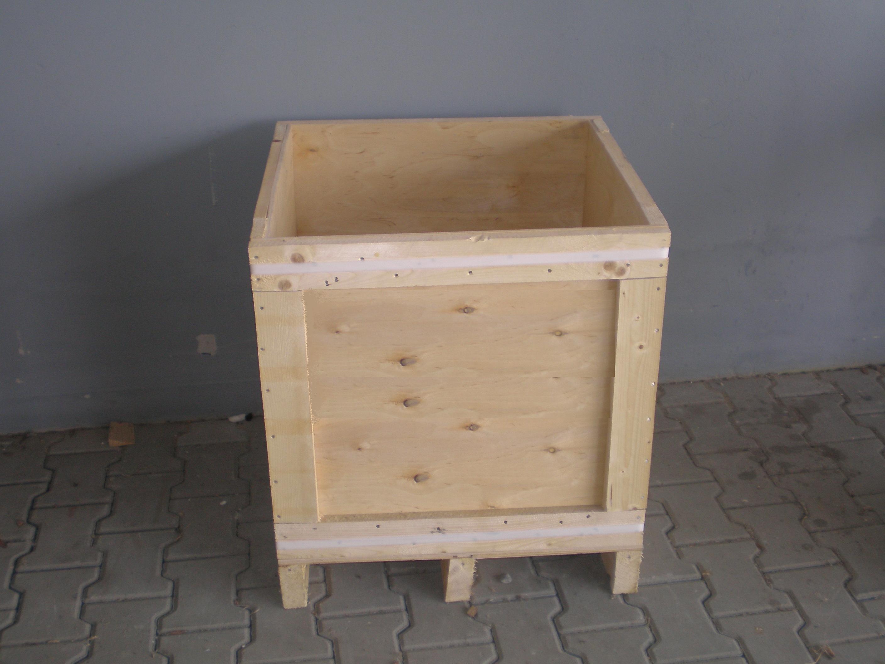 Pusta, jasna, drewniana skrzynia paletowa z czterema nogami, zabezpieczona taśmą, stojąca na szarej kostce brukowej przed szarą ścianą.