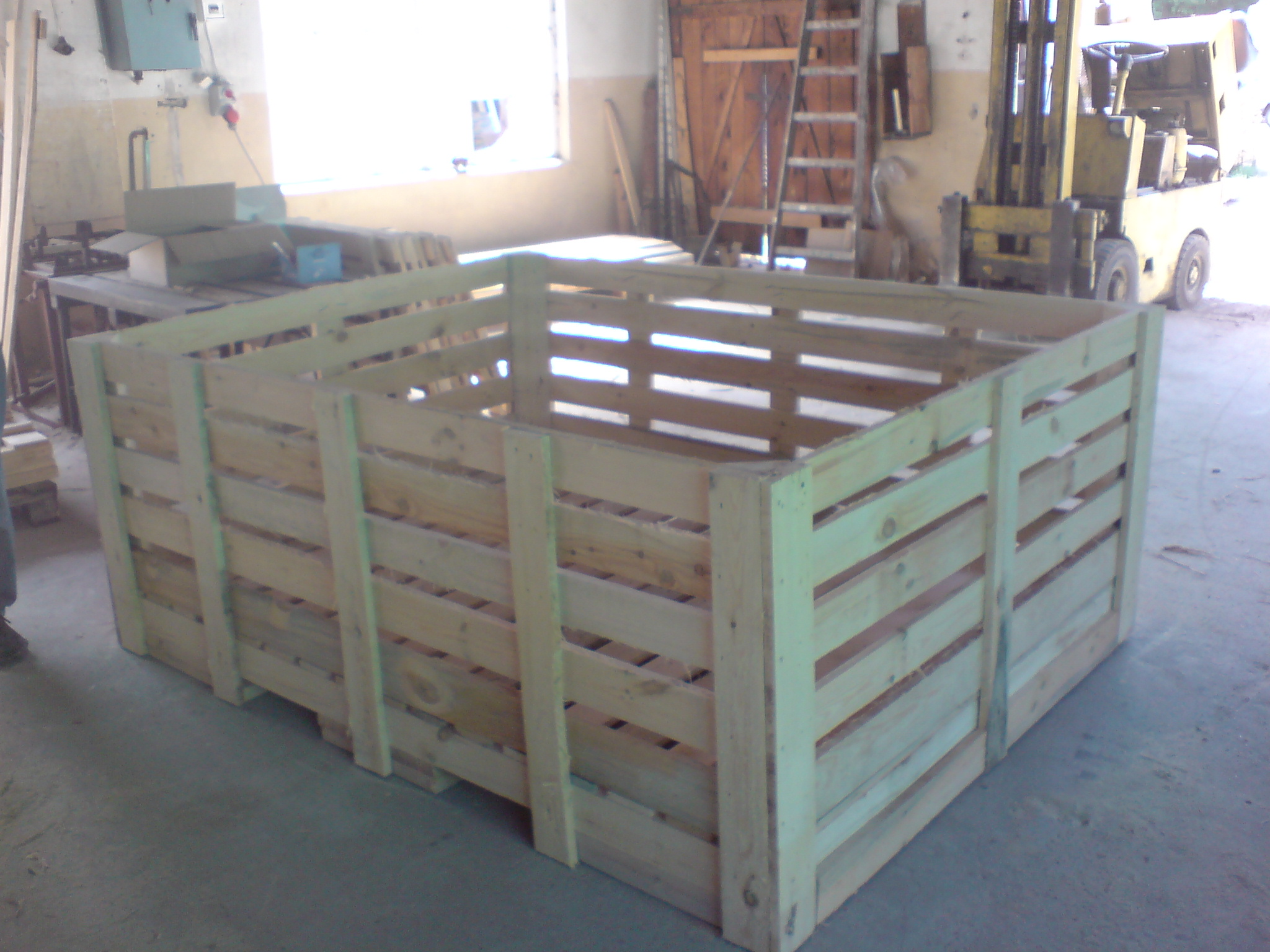 Duża, pusta skrzyniopaleta z jasnego drewna, widoczna w hali produkcyjnej z wózkiem widłowym w tle.