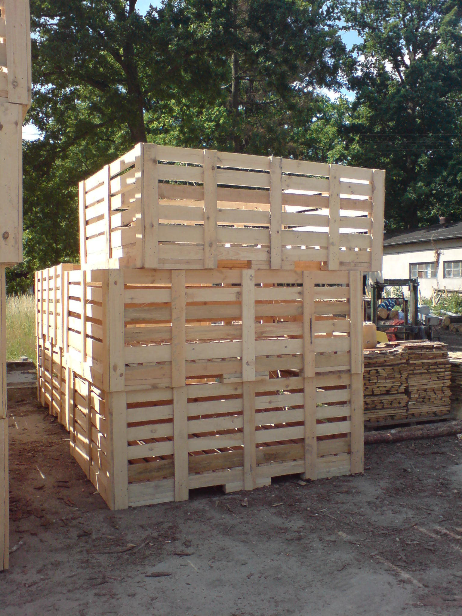 Stos jasnych, drewnianych skrzyniopalet ustawionych na zewnątrz, z widocznymi deskami i konstrukcją kratową, tło z drzewami i budynkiem, w oddali wózek widłowy i ułożone deski.