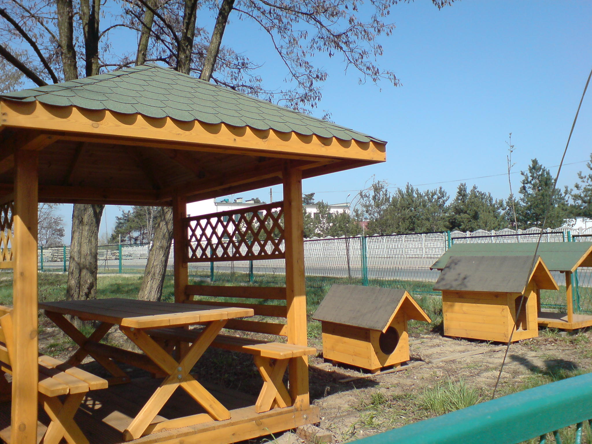 Drewniana altana z zielonym dachem, stół z ławkami wewnątrz, obok dwie budy dla psów z drewna, w tle drzewa i niebieskie niebo.