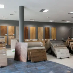 Ekspozycja paneli podłogowych w sklepie: różnorodne wzory i kolory ułożone na stojakach, pionowe panele na ścianie, widoczny fragment podłogi z płytek imitujących drewno.