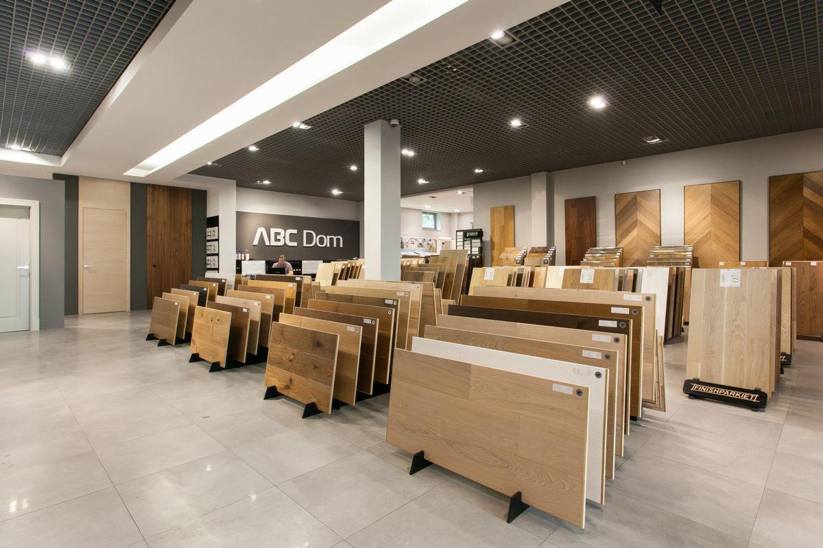 Ekspozycja paneli podłogowych w sklepie ABC Dom: różnorodność wzorów i odcieni drewna prezentowana na stojakach, z widocznym pracownikiem za ladą i próbkami drzwi w tle.