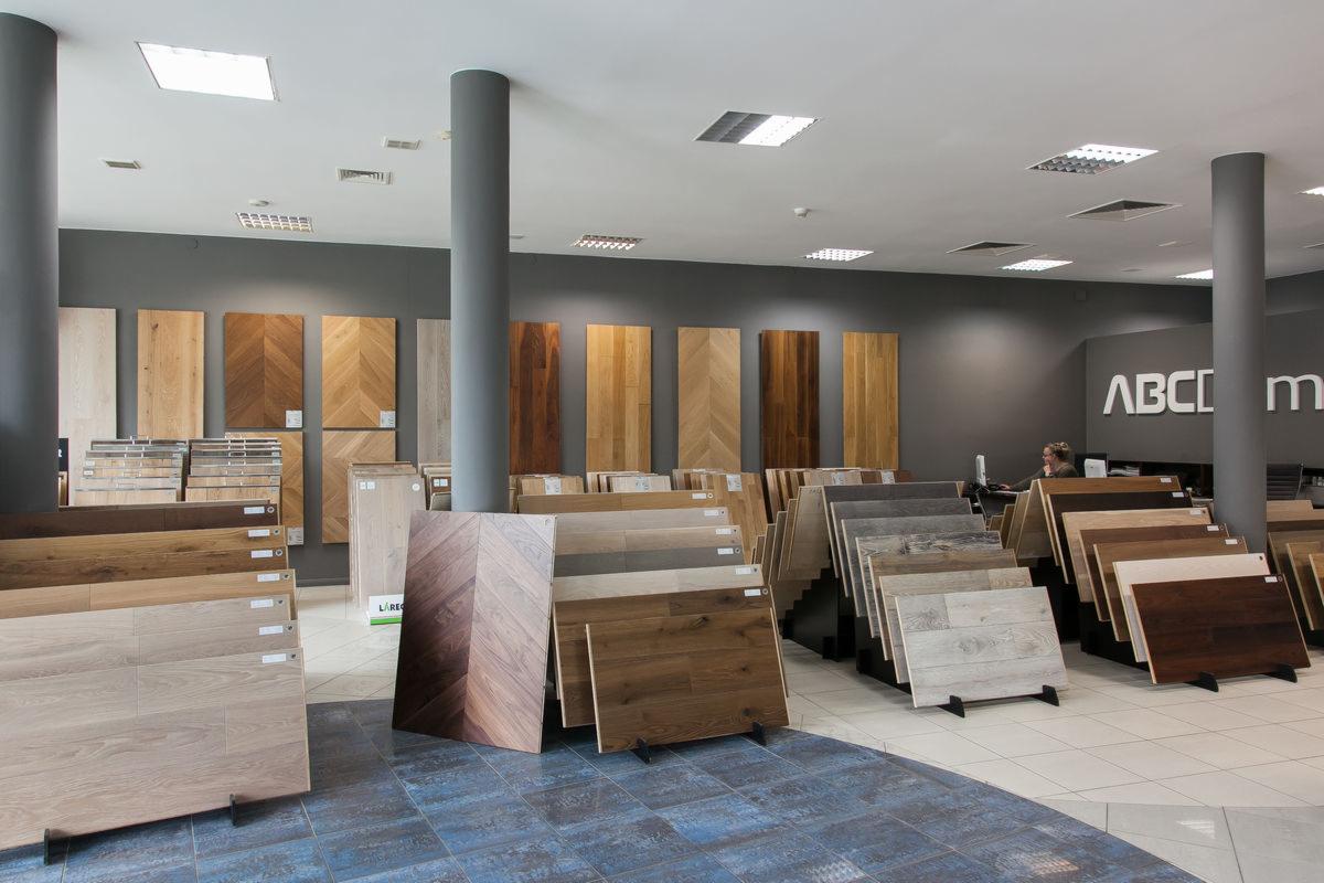 Ekspozycja paneli podłogowych w sklepie: różnorodne wzory i kolory ułożone na stojakach, pionowe panele na ścianie, widoczny fragment podłogi z płytek imitujących drewno.