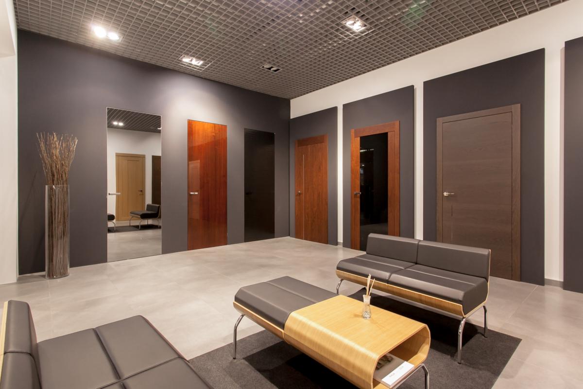 Wnętrze salonu z ekspozycją różnych modeli drzwi wewnętrznych w odcieniach brązu, ustawionych w ciemnoszarych wnękach, z nowoczesnymi ławkami dla klientów i dekoracyjnym wazonem z suchymi gałązkami.