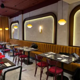 Wnętrze restauracji z rzędami stolików i krzeseł w odcieniach czerwieni i brązu, oświetlone wiszącymi lampami w stylu retro i ścianami z dekoracyjnymi wnękami.