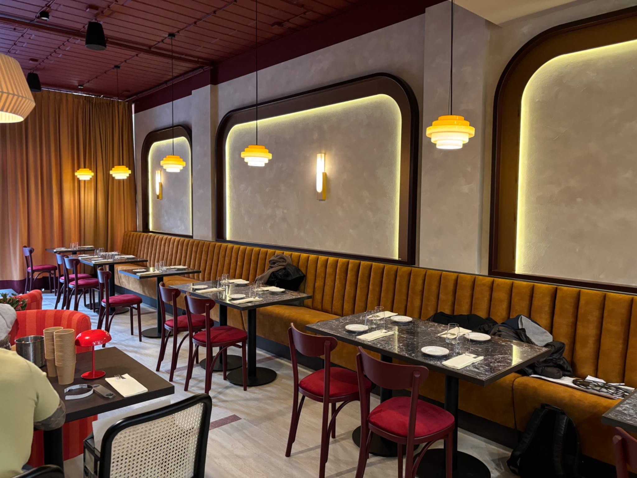 Wnętrze restauracji z rzędami stolików i krzeseł w odcieniach czerwieni i brązu, oświetlone wiszącymi lampami w stylu retro i ścianami z dekoracyjnymi wnękami.