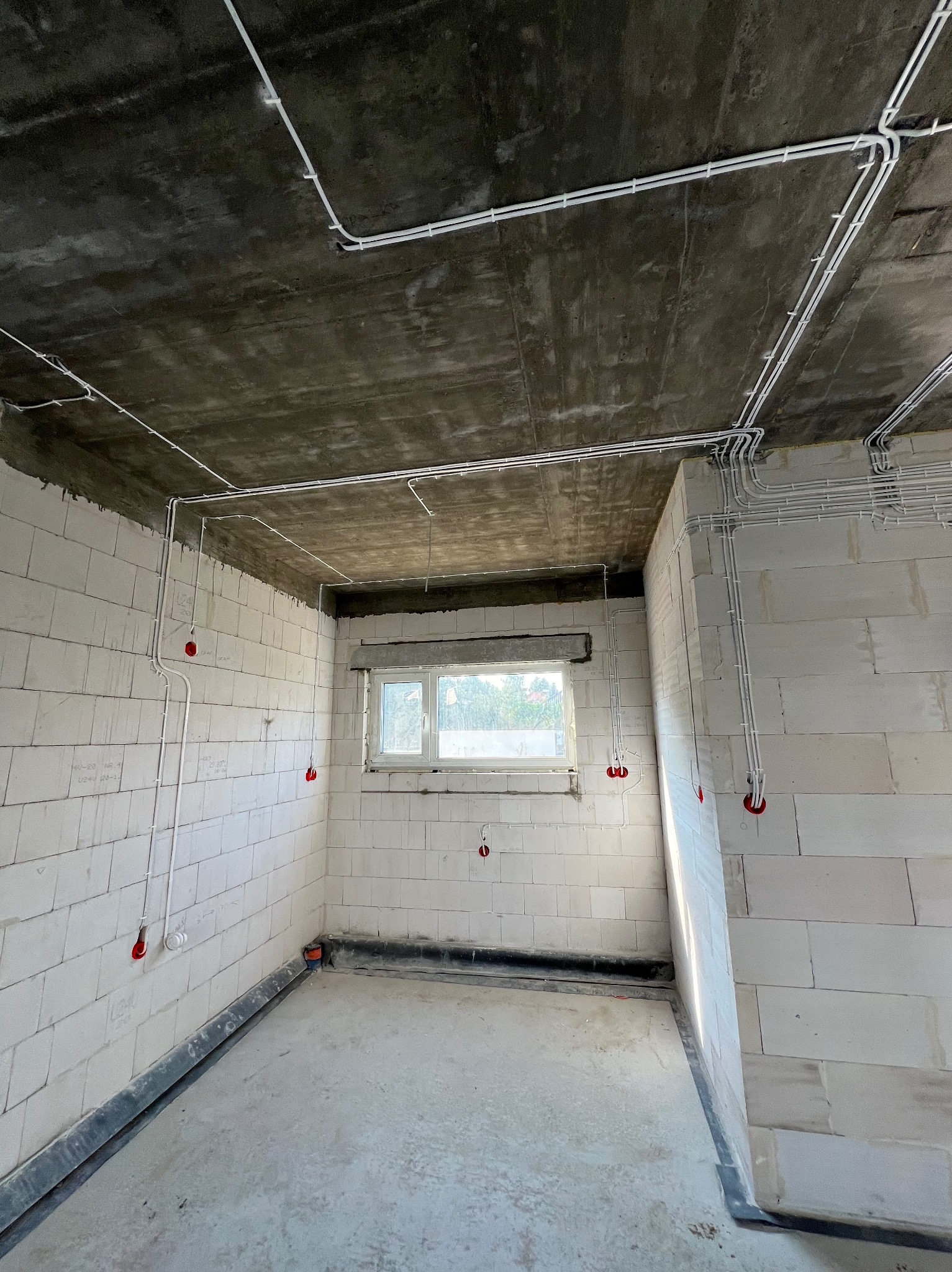 Surowy stan: białe ściany z betonu komórkowego i szary betonowy sufit z widoczną instalacją elektryczną w białych peszlach i czerwonymi puszkami w nieukończonym wnętrzu.