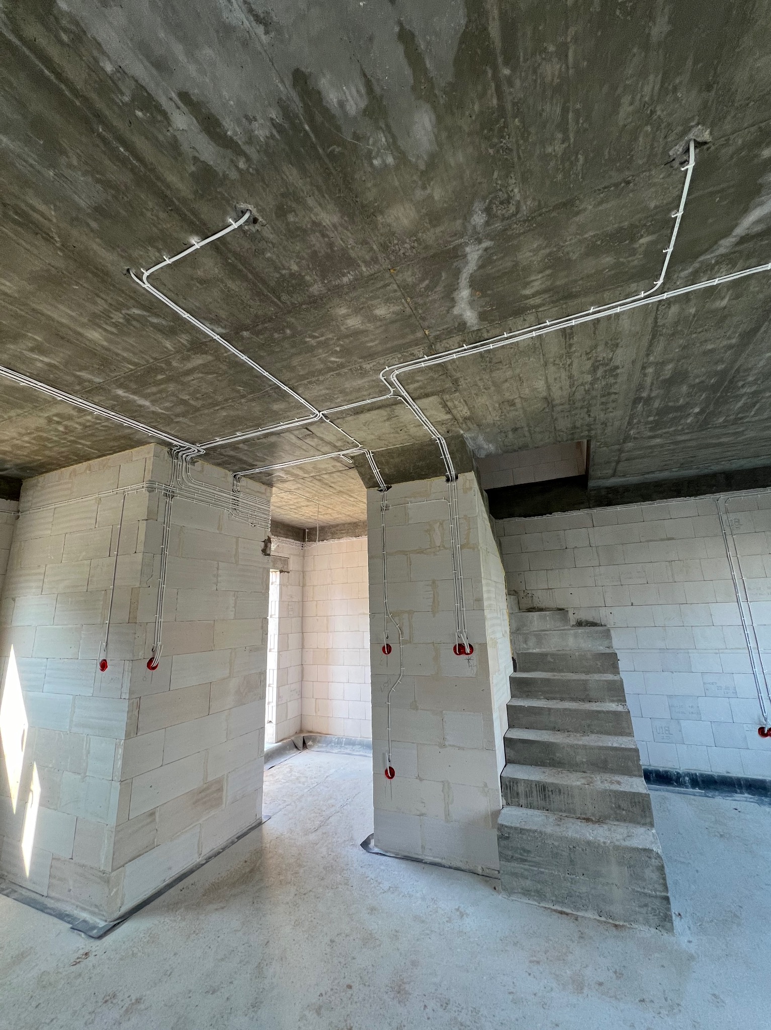 Surowy stan wnętrza z instalacją elektryczną: białe kable ułożone na ścianach z betonu komórkowego i suficie, widoczne czerwone puszki instalacyjne, betonowe schody