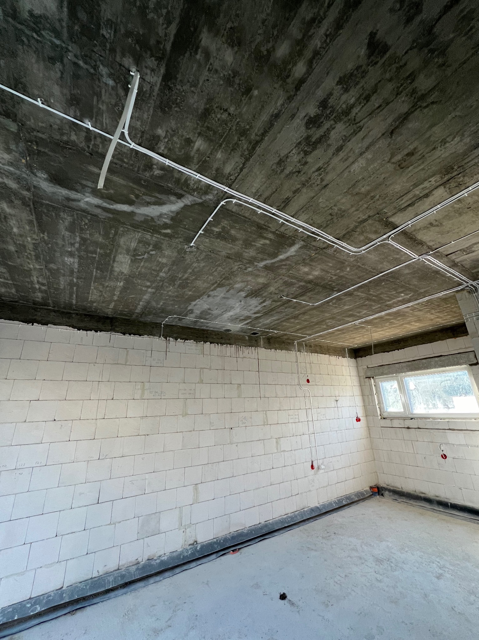 Surowy pokój z białymi ścianami z betonu komórkowego i widocznymi przewodami elektrycznymi poprowadzonymi po suficie i ścianach, zakończonymi czerwonymi puszkami instalacyjnymi.